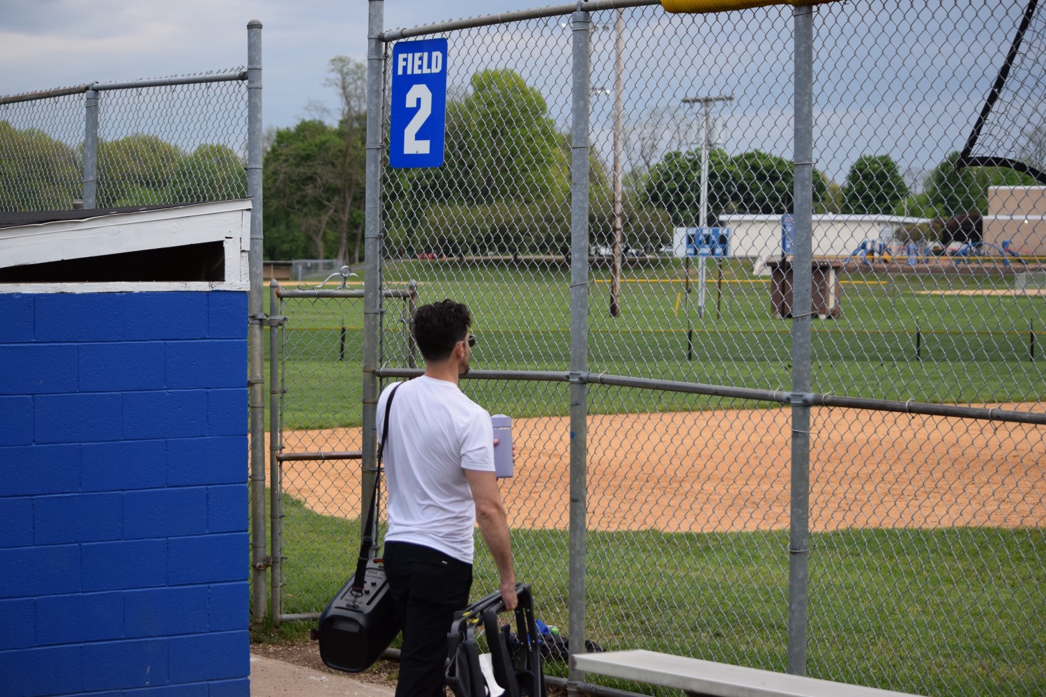 Andrew walking up to Field 2 — folding chair and speaker in hand, looking out at the empty diamond.